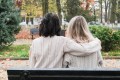 Back view of two women sitting on a bench, one supporting the other with her arm over her