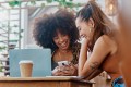 Two women sitting at a coffee shop, laughing and looking at a phone