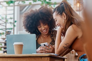 Two women sitting at a coffee shop, laughing and looking at a phone